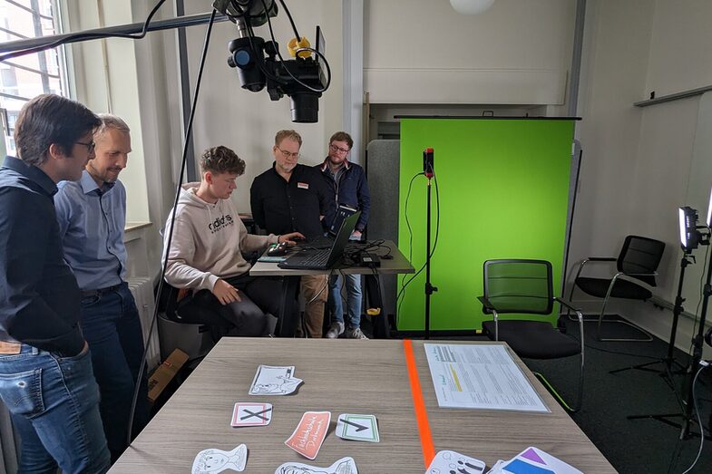 In one room there is a green screen in the background and a table in the foreground, on which are cut-out drawings of symbols. A camera is mounted above the table. Five people have gathered around a laptop.