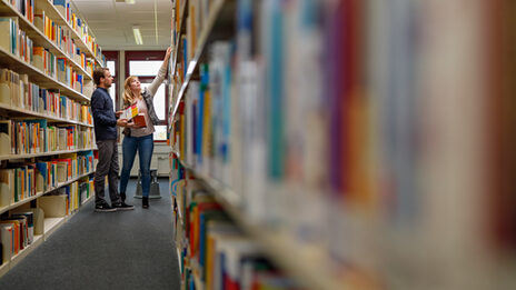Photo of a female and male student standing in a row of shelves in the Emil-Figge-Strasse library, fetching books from the shelf __A female and male student stand in a row of shelves in the Emil-Figge-Strasse library, fetching books from the shelf.