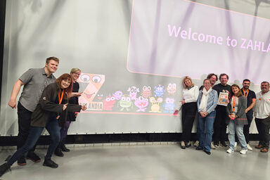 Zehn Personen stehen vor einer Leinwand, auf die "Welcome to ZAHLANDIA" und eine Reihe bunter Monsterfiguren projiziert ist.