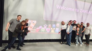 Ten people stand in front of a screen on which "Welcome to ZAHLANDIA" and a series of colorful monster figures are projected.
