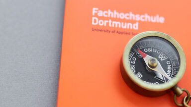 Foto eines kleinen Kompasses mit Kette daran, der mit einem Bleistift auf einer orangefarbenen Mappe der Fachhochschule Dortmund liegt.