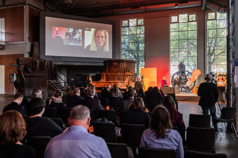 Publikum sitzt vor einer Bühne, auf der drei Menschen zu sehen sind. Links von der Bühne ist eine große Leinwand, auf der zum einen die Bühne in Nahaufnahme als auch eine hyprid zugeschaltete Person zu sehen sind.