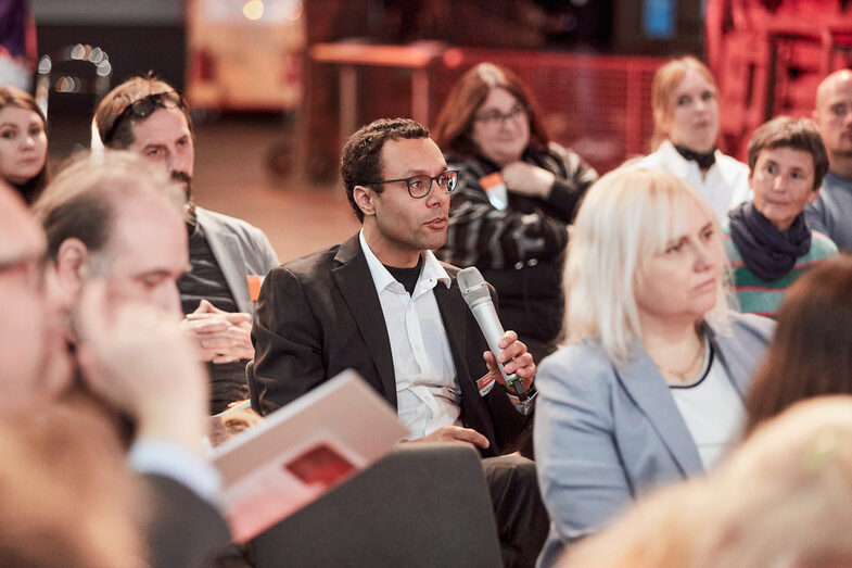 Lehrender im Publikum stellt eine Frage an Teilnehmende einer Podiumsdiskussion.