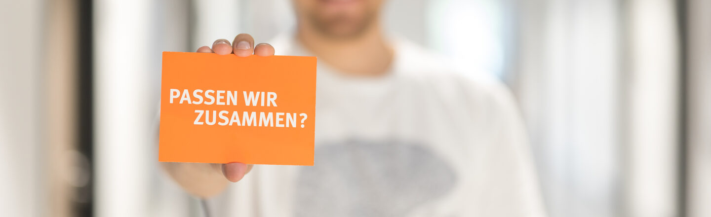 Foto eines Mannes, der eine Karte mit Schriftzug "Passen wir zusammen?" in die Kamera hält.__Man out of focus holding a card with the words "Do we fit together?" into the camera.