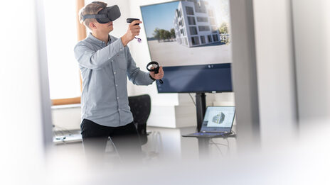 Foto von einem Mann mit VR-Brille auf und Controller in der Hand. Auf dem Bildschirm im Hintergrund sieht man ein Gebäude in der Perspektive, die auch der Mann durch die VR-Brille sieht.