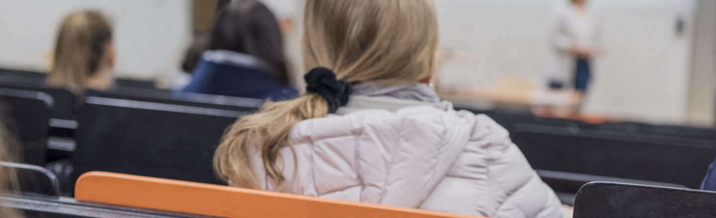 Foto einer jungen Frau in Rückansicht in einer Hörsaalreihe, diet nach vorne zur vortragenden Person schaut. In der Reihe dahinter ist der Tisch aufgeklappt, darauf liegen Printmaterialien. __ <br>Young woman in a row of lecture halls, looks forward to the speaker. In the row behind it, the table has been opened, and there are print materials on it.