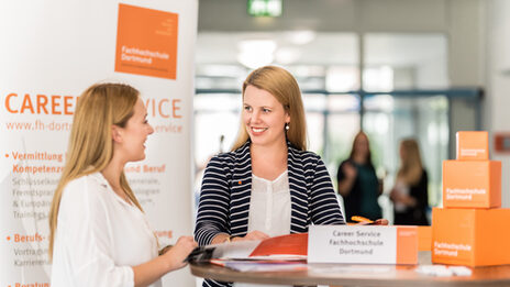 Photo of an employee of the Career Services of the Fachhochschule Dortmund standing at a high table talking to a young woman. __ <br>Employee of the Career Services of the University of Applied Sciences Dortmund stands at a high table talking to a young woman.