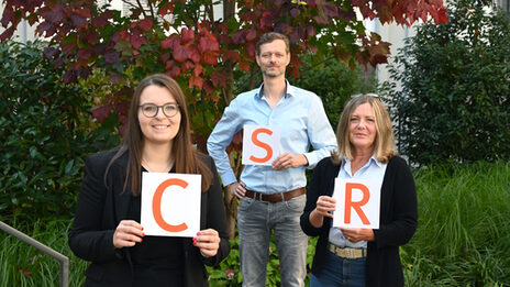 Group picture of CSR Office teams: three people each hold up a sheet of paper with an orange letter on it. From left to right, this is how CSR is read.
