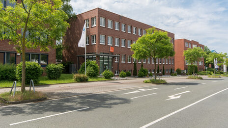 Photo of the Fachhochschule Dortmund site in Otto-Hahn-Straße from the outside. It is a red brick building. In the foreground are a road and some trees.