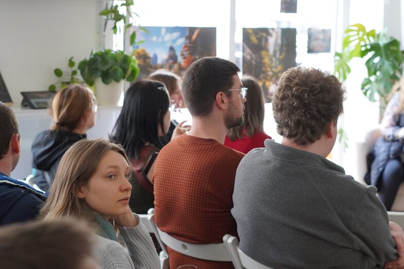 While the audience looks ahead, a member of the audience turns her head to the side. There are tall plants scattered around the room.