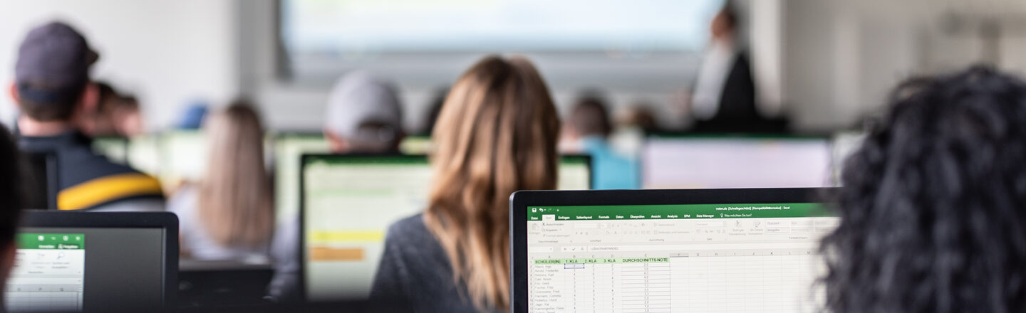 View from behind into the computer room with students, some Excel spreadsheets are visible on the monitors.