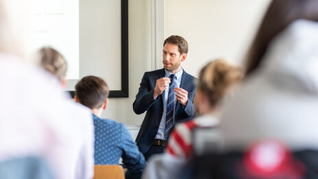 Foto eines Lehrenden, der vor seinen Studierenden steht und etwas erläutert