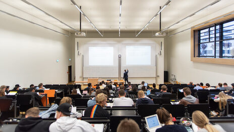 View of a full lecture hall from the back, a lecturer is writing something on the blackboard at the front.