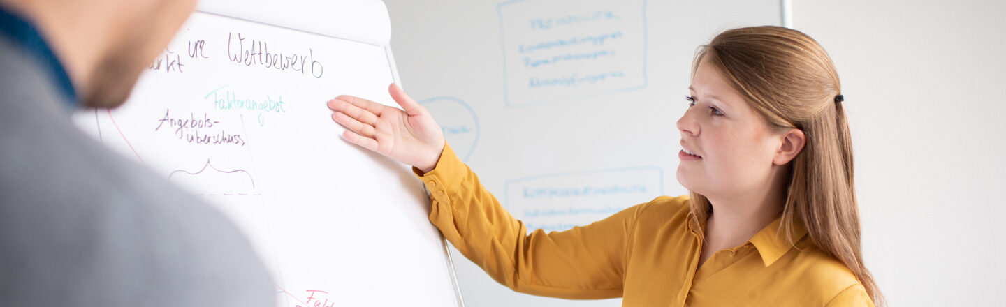 Studentin zeigt auf das Wort "Wettbewerb" auf einem Flipchart und erläutert ihrem Kommilitonen gegenüber etwas.