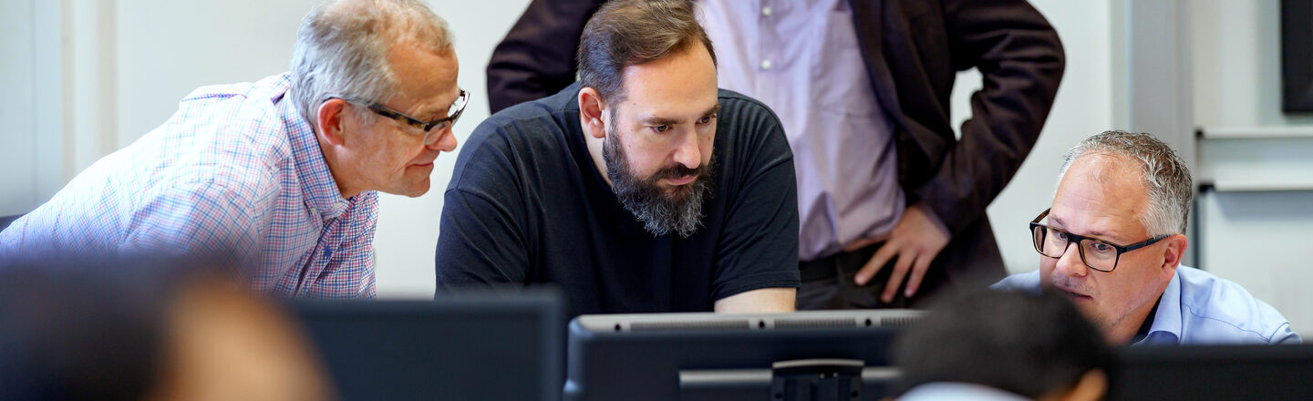 Photo of four lecturers looking into a laptop at the front of the room. Students are sitting in the foreground, out of focus.