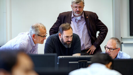 Photo of four lecturers looking into a laptop at the front of the room. Students are sitting in the foreground, out of focus.