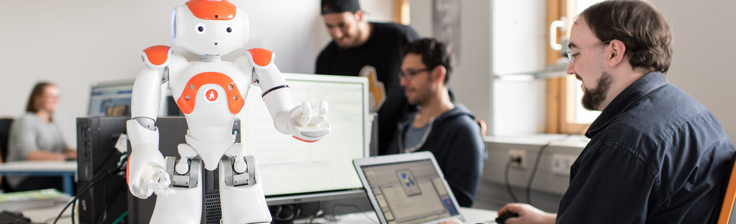 Photo of the NAO robot Eva standing on the table in front, arms raised. Next to it, a student sits at the laptop to give Eva commands.