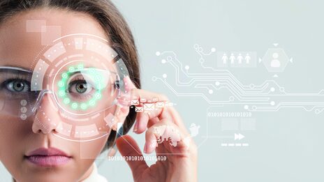 Portrait photo of a woman wearing glasses with a button on the side that she presses. Circles and shapes are animated around her left eye, with other symbols to her left.