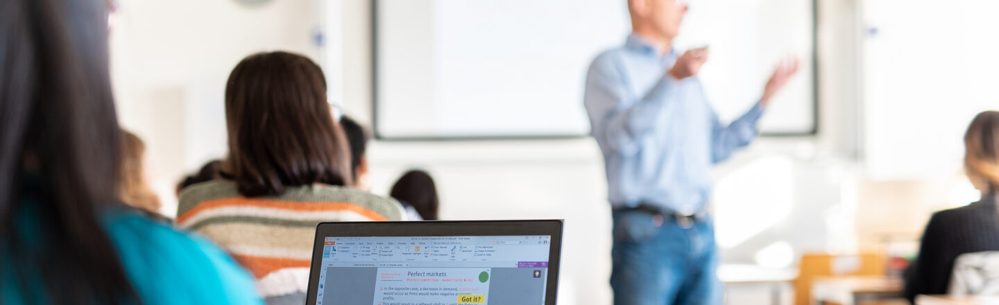 View of a student's laptop in a row of seats. Blurred in front of her, a lecturer explaining something.