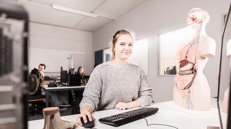 Foto einer Studentin an einem Rechnerarbeitsplatz, neben ihr auf dem Tisch steht ein Anatomiemodell.