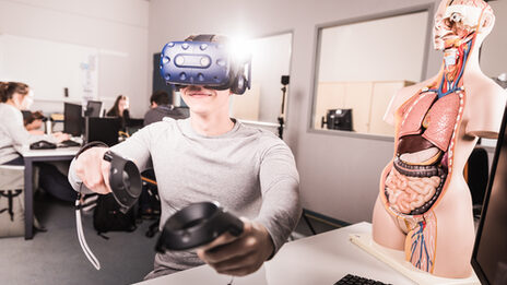 Foto von einem Studenten, der eine VR-Brille auf hat und Controller in der Hand hält, er sitzt an einem Rechnerarbeitsplatz.__A student has VR glasses on and controller in his hands, is sitting at a computer workstation.