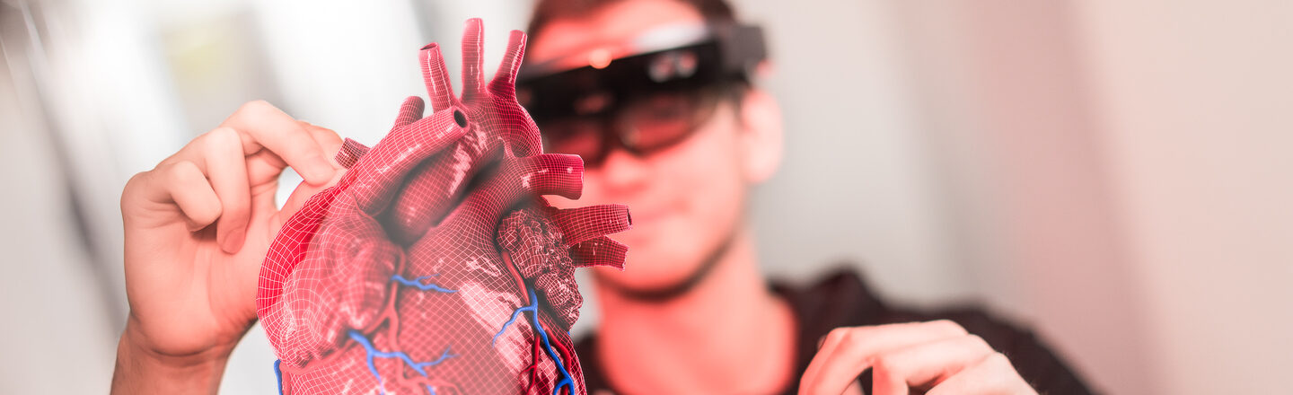 Foto von einem Studierenden mit VR-Brille, der die Hände zur Steuerung in die Höhe hält. Vor ihm sieht man, was er durch die Brille sieht: ein 3D-Herz-Hollogramm. __ Student wears VR glasses and holds hands up to control. In front of him you can see what he sees through the glasses: a 3D heart hollogram.