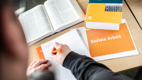 Photo of someone writing something on a pad with an orange pen. Next to it are books and a notebook labeled "social work".