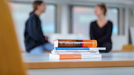 Photo of a pile of books on a table. Two people talking can be seen out of focus in the background.