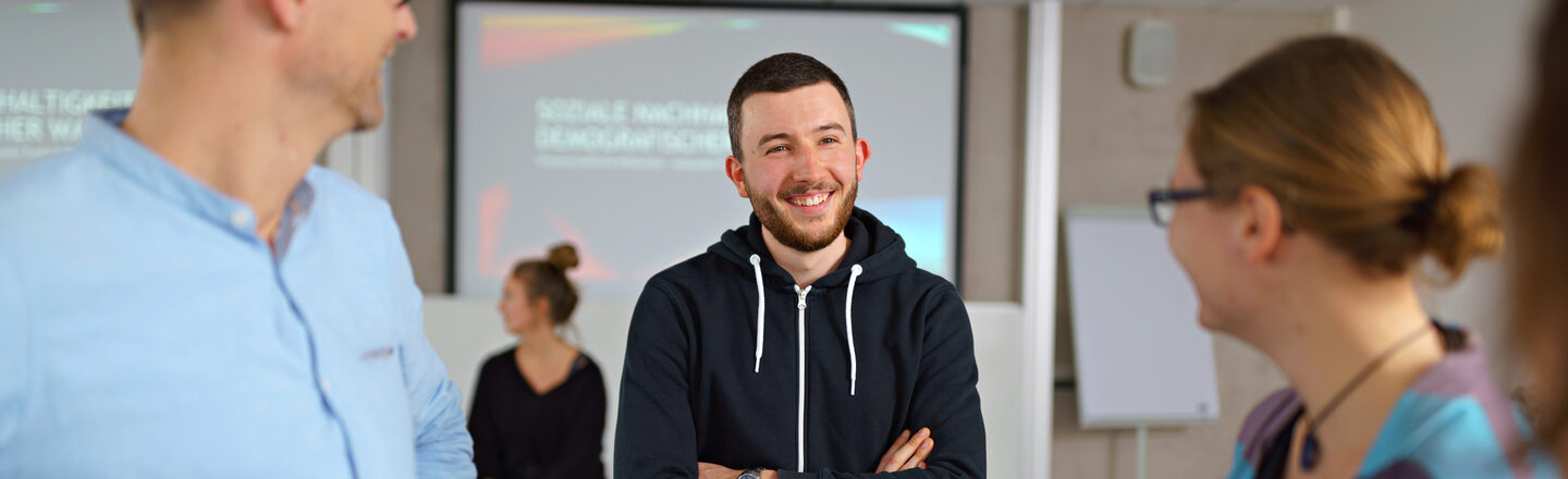 Foto von drei Personen, die beieinander in einem Seminarraum stehen und sich anlachen.