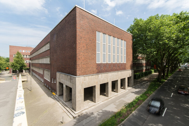 Foto mit Blick auf Teile eines Gebäudes der Fachhochschule Dortmund am Max-Ophüls-Platz mit Vorplatz und Gehweg sowie Straße mit Autos daneben.