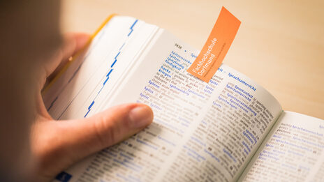 Photo of an open dictionary held in someone's hand. The open page is marked with an orange slip of paper with the word mark of Fachhochschule Dortmund __Photo of an open dictionary held in someone's hand. The open page is marked with an orange slip of paper with the word mark of Fachhochschule Dortmund.