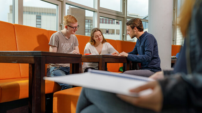 Photo of a female student and two male students sitting on orange chairs. They are looking together at documents on the table. On the right in the picture another student in the cut.__One female and two male students on orange chairs, they look together at documents on the table. On the right in the picture another student in the cut.