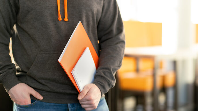 Photo of a student - head down - who wears a sweater with the logo of the Fachhochschule Dortmund and holds some folders and pads in his hand. __ A student wears a sweater with the logo of the Dortmund University of Applied Sciences and holds some folders and pads in his hand.