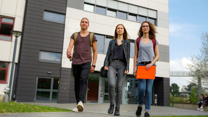 Photo of two female and one male student walking side by side on a path, in the background a building on Emil-Figge-Straße and other students __Two students walk side by side on a path, in the background a building on Emil-Figge-Straße and other students.