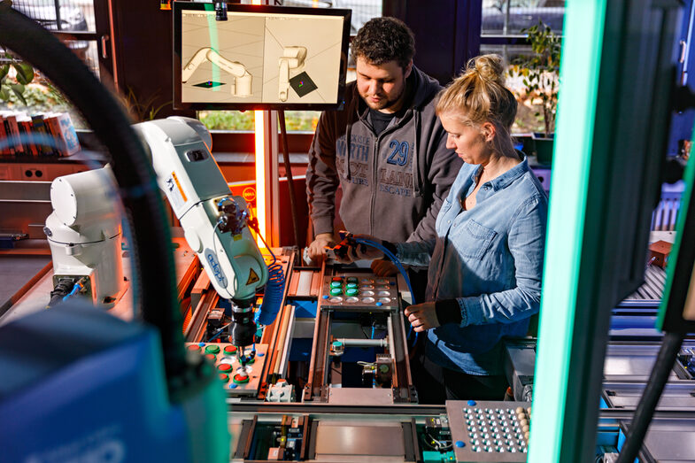 Foto von zwei Personen, die etwas an einem Förderband mit zugehörigem Roboterarm einstellen.