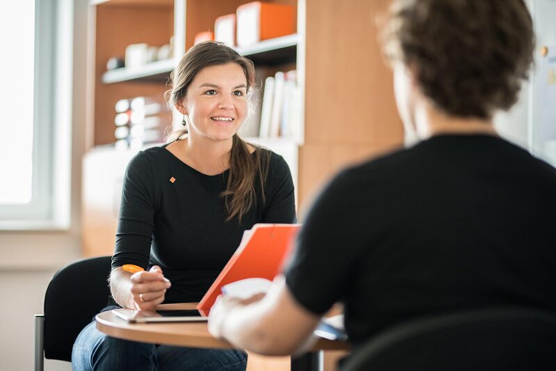 Foto von einer Mitarbeiterin der Studienberatung, die einem Studenten gegenüber sitzt, den man in Rückansicht sieht. Sie führen gemeinsam ein Beratungsgespräch.