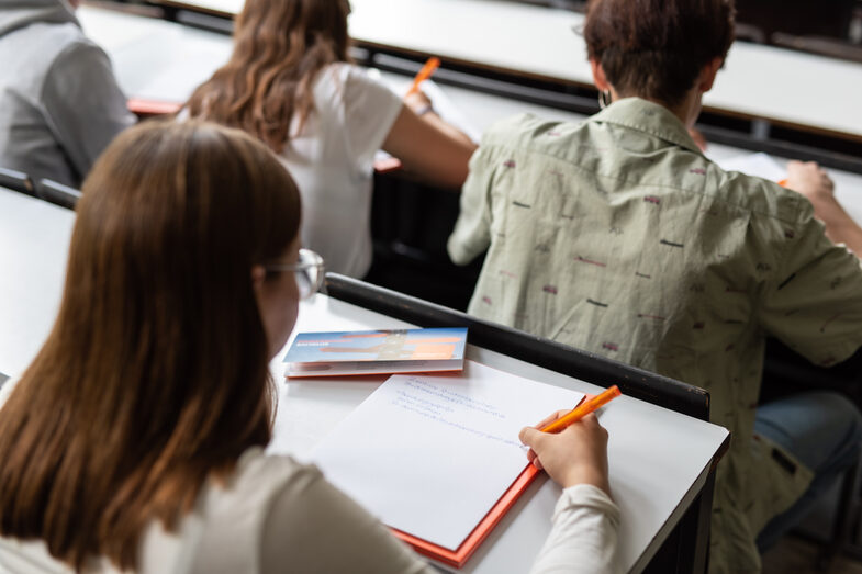 Foto einer jungen Frau, die sich Notizen im Hörsaal macht. Vor ihr sitzen drei weitere Personen. __ Young woman takes notes in the lecture hall, three other people are seated in front of her.