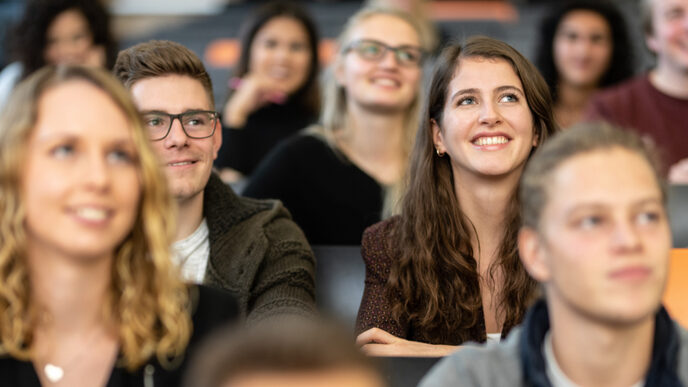 Close-up of several students in the rows of seats in a lecture hall. The focus is on a female student and a male student sitting next to each other and looking straight up.