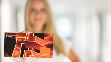Photo of a postcard from the student advisory service, on which illuminated stairs and the words "Master's course offer" are printed. A woman holds the card up to the camera.