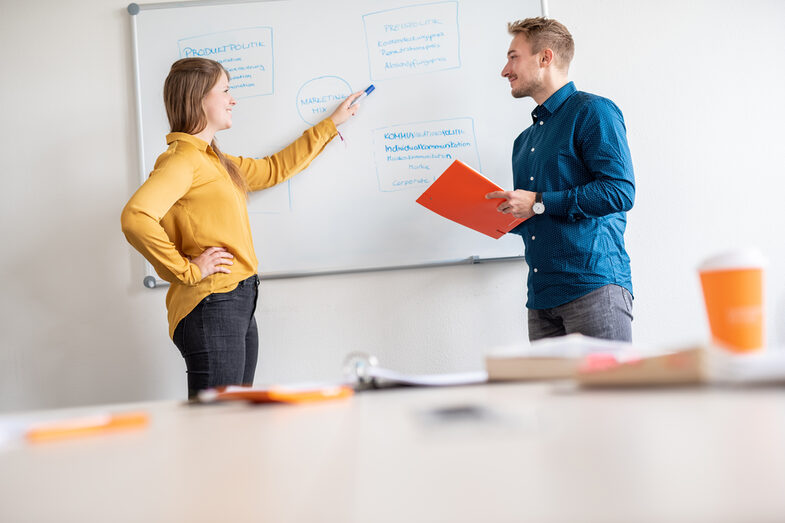 Zwei Studierende stehen vor einem Board, auf dem eine Grafik zum "Marketing-Mix" aufgezeichnet ist. Die Studentin links im Bild, zeigt dort auf eine "Box" und schaut dabei Ihren Kommilitonen an.