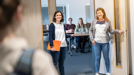 Photo of a student standing on the left of the picture with her back to the camera. She is looking towards the door opposite her, where two members of staff are standing and greeting her warmly. A seminar room with other people is blurred in the background.