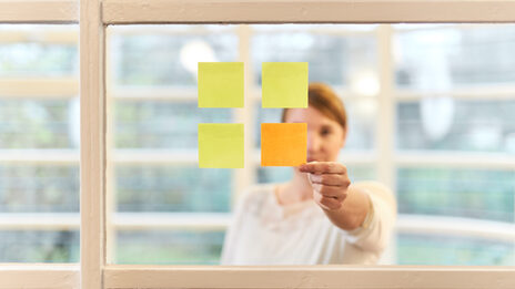 Frontal view of a woman standing behind a pane with four square Post-its stuck to it, which in turn form a square. The woman grabs the only orange Post-it.
