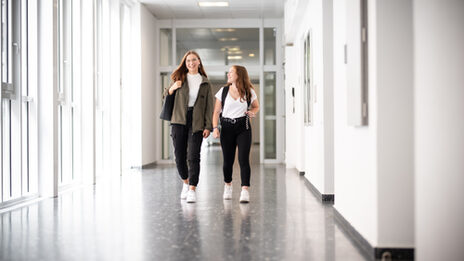 Foto von zwei Schülerinnen, die lachend über den Gang laufen. __ Two girls run down the hallway laughing.