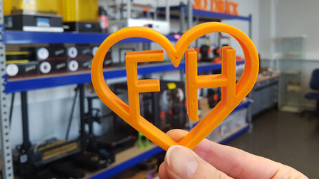 One hand holds an orange heart from the 3D printer, which encloses the letters F and H. In the background are shelves with appliances.