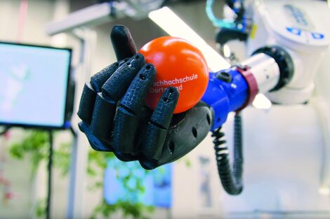 Nahaufnahme einer bionischen Hand an einem Industrieroboter, die einen orangefarbenen Ball mit der Aufschrift "Fachhochschule Dortmund" hält.