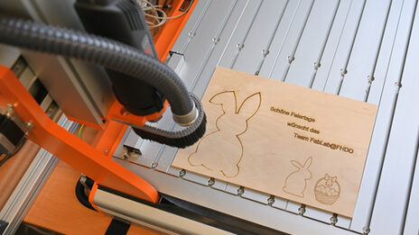 On a laser cutter lies a thin wooden plate with the outlines of two rabbits, a basket with eggs and the sentence: Happy holidays from the FabLab@FHDO team.