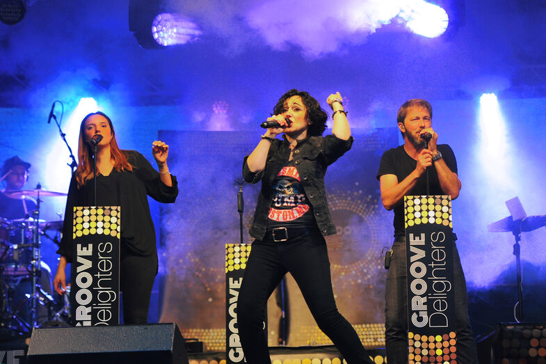 Two female and one male singer stand on an illuminated and fogged stage and each sing into a microphone. The microphone stands are labeled with the words Groove Delighters.