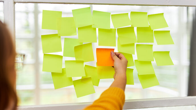 Photo of many yellow Post-its stuck next to and below each other on a window pane. A single orange Post-it is stuck in the middle. A woman stands in front of it and reaches for the orange Post-it.