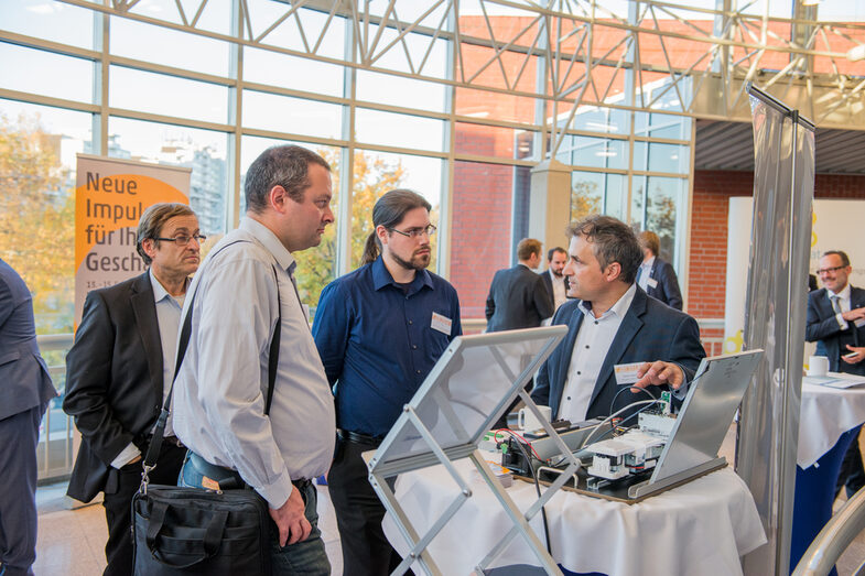 Photo from the SMARTenergy conference: A man explains a technical product on display to three men at a high table.