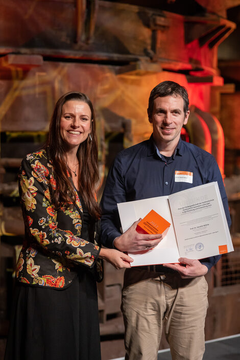 Photo of a male and a female person standing and smiling at the camera. The male person is holding a dice and a certificate.